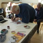 1 elderly man creating artwork as 2 others chat